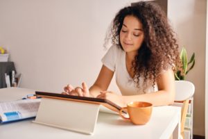 Smiling girl with curly hair sitting at the table with book and cup of coffee dreamily using tablet